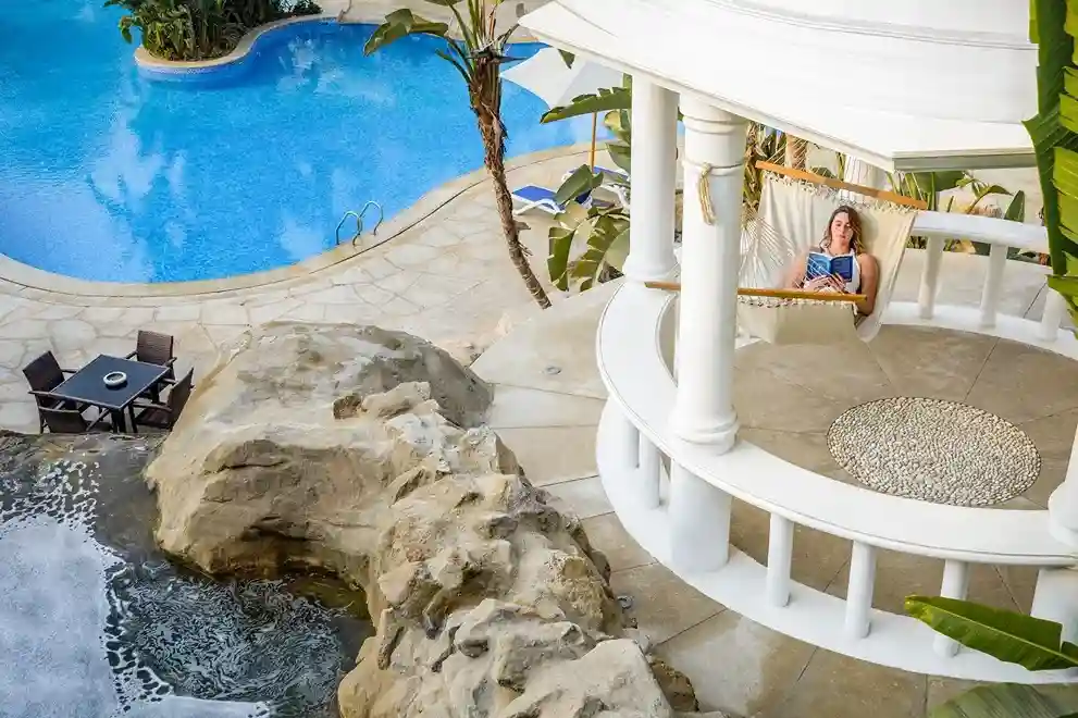 A woman relaxes in a hammock on a white balcony overlooking a blue swimming pool and palm trees; a table with chairs is set up nearby on a stone patio.